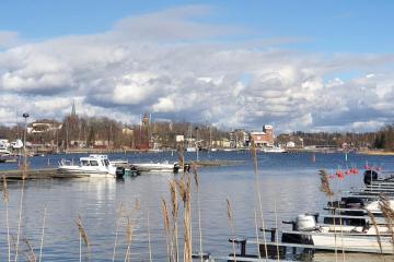 Kaunis, huhtikuinen kuva Kaupunginlahden alkupäästä, jossa näkyy edessä kaisloja, niiden takana laitureissaan olevia veneitä, ja taaempana näkyy kaupunki.
